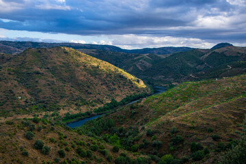 Naklejka premium A la izquierda España, a la derecha Portugal: Frontera entre ambos países marcada por el río Duero en una imagen al atardecer con vegetación verde y nubes oscuras