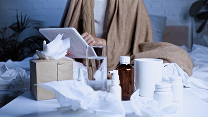cropped view of sick woman using digital tablet near medications, drinks and paper napkins on blurred foreground