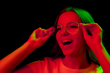 Caucasian woman's portrait isolated on black studio background in multicolored neon light. Puts on the eyeglasses. Concept of human emotions, facial expression, sales, ad, fashion. Copyspace.