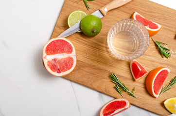 Top view of glass full of mineral water, slices of grapefruit, rosemary, lemon and lime on the wooden board, white table