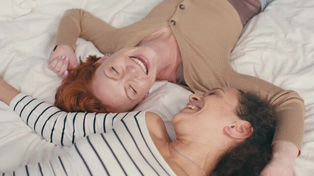 high angle view of pleased interracial lesbian couple laughing and lying on bed