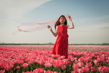 Teenage girl standing in a field of pink tulips wearing a red dress holding thin red  fabric in the wind