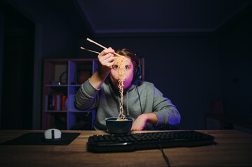 Funny gamer has dinner with a lamp at the computer at night in a room with purple light. Guy in a...
