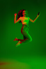 Fototapeta na wymiar Jumps high. Caucasian woman's portrait on green studio background in multicolored neon light. Beautiful female model. Concept of human emotions, facial expression, sales, ad, fashion. Copyspace.