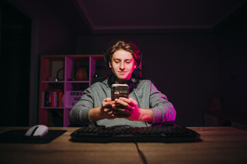 Handsome guy is sitting at the computer at night and uses a smartphone with a smile on his face.Gamer with a headset around his neck uses the Internet on phone and smiles in room with a purple light.