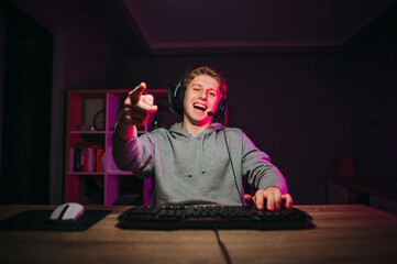Happy young male gamer in headset and casual makes streaming game and communicates with subscribers and shows finger to camera and smiles. © bodnarphoto