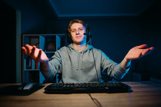 Positive young male gamer in a headset on his head spreads his arms to the sides and looks at the screen with a smile on his face. Guy stream game online at night in the room