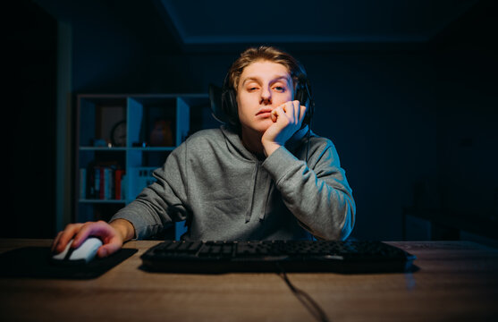 Tired young man in headset playing video games at home on computer in room with blue light and looking at camera with sleepy face.
