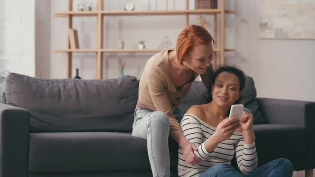 multicultural lesbian couple looking at smartphone and smiling in living room