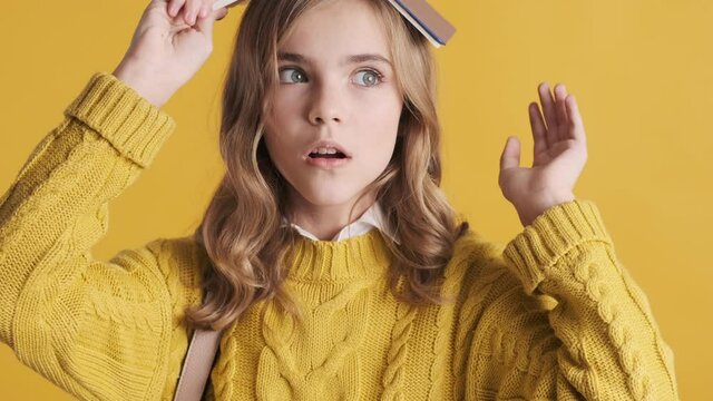 Pretty blond teenage student girl posing with book on head looking happy over yellow background. Funny pupil girl fooling around on break