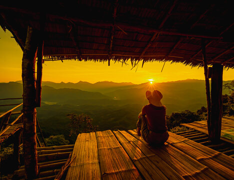 Picture from the back of a woman sitting on wooden porch extending into a high mountain cliff. The sun is setting on the mountain and there is a beautiful warm orange light. The traveling background.