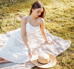 Girl with a straw hat in the spring in the park. Brunette with long hair sitting on a plaid on a background of summer nature. Youth and beauty.