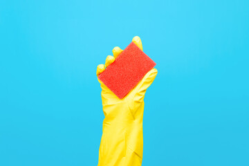 Housewife's hand in yellow rubber glove holding red washing sponge, blue background. Household cleaning equipment concept