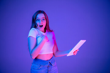 Caucasian woman's portrait isolated on blue studio background in multicolored neon light. Beautiful model with tablet. Concept of human emotions, facial expression, sales, ad, fashion. Copyspace.