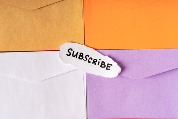 A white piece of paper with the inscription SUBSCRIBE is lying in a pile of colorful envelopes. Bright background