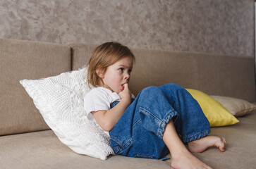 a little girl in a blue denim jumpsuit sits on the couch and bites her nails.
