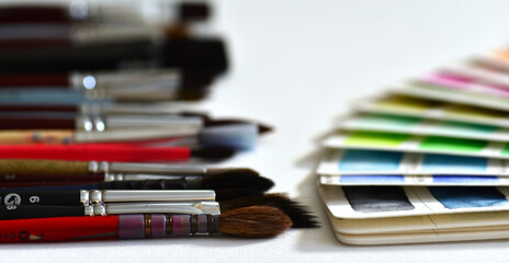 Watercolor color palette on white paper, fanned out, next to a set of brushes. On a white paper background. Fragment close-up.