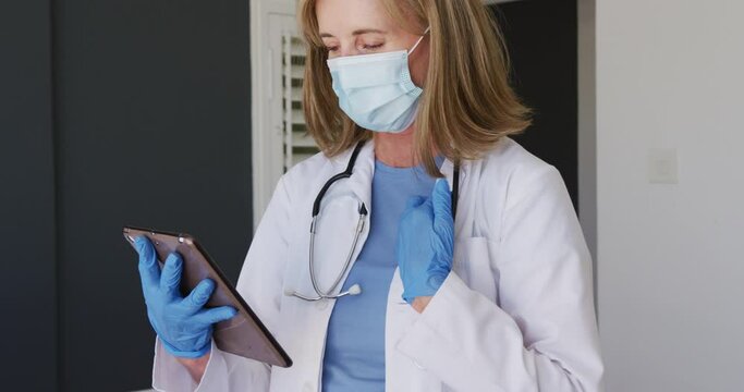 Caucasian senior female doctor wearing surgical gloves and face mask using tablet computer
