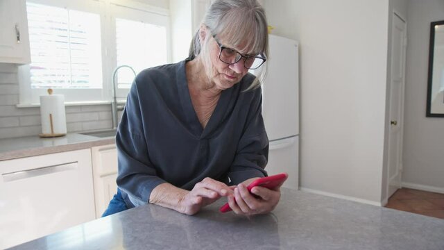 Senior Caucasian woman text messaging on smartphone while sitting at kitchen counter. Older white lady working on cell phone at home. Slow motion 4k