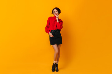 Fototapeta na wymiar Good-looking girl in red sweater standing on yellow background. Studio shot of joyful lady with short hair wears skirt.