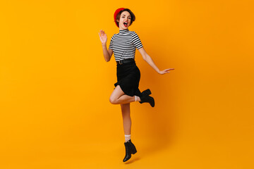 Carefree french woman in skirt dancing on yellow background. Studio shot of jumping female model in red beret.