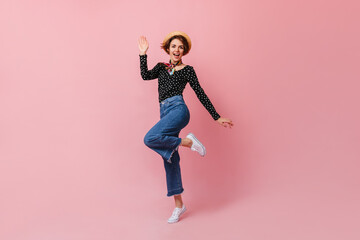 Wonderful young lady in straw hat jumping on pink background. Studio shot of girl in denim pants dancing with smile.