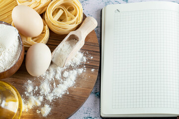 Tagliatelle with oil, egg and bowl of flour on wooden board and notebook