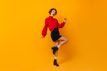 Inspired woman in short skirt dancing on yellow background. Studio shot of blissful brunette girl in red sweater.