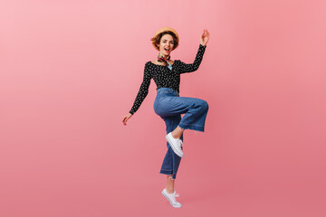 Fototapeta na wymiar Full length view of jumping woman in straw hat. Studio shot of girl in vintage jeans dancing on pink background.