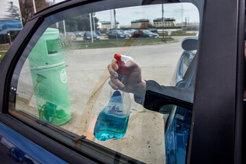 Mano masculina sosteniendo un spray limpiacristales cerca de una ventana de coche con la puerta...