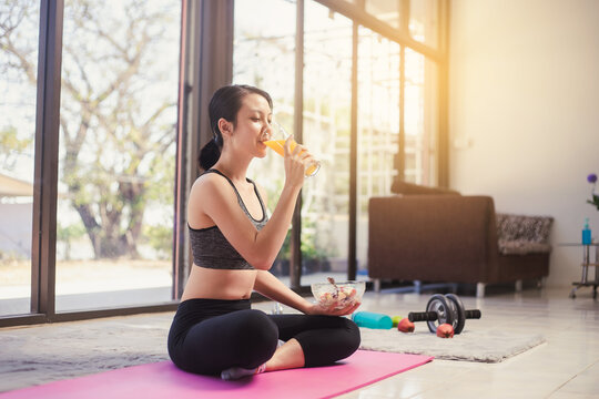 Healthy Asian Woman In Sportswear Sitting On The Yoga Mat Drinking Orange Juice And Eating Salad Fruit At Home. Eating Diet And Exercise Relaxation Concept.