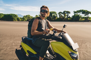 Cool young man riding motorcycle and using smartphone on sandy shore of tropical country