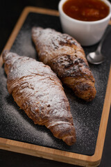 Blurred image of croissants and apricot jam in a gravy boat on a board against a dark background.