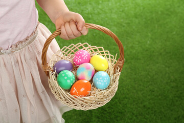 Little girl with basket full of Easter eggs on green grass, closeup. Space for text