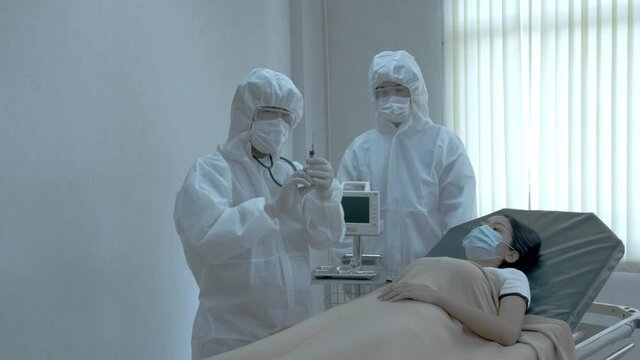 Medium Shot – An Asian Male Doctor Wearing PPE Suit Giving An Injection To Female Patient Affected By Coronavirus Or COVID 19 In A Hospital.