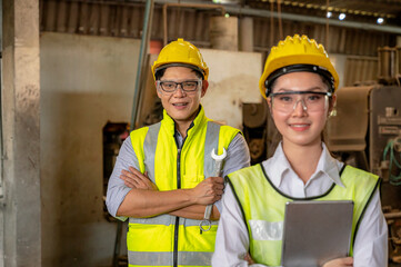 Nonthaburi - Thailand 06 February 2021 : Female and male Asian engineers smiling, wearing hard helmets and holding tablets, both looking at the camera standing at the machinery area in the factory are