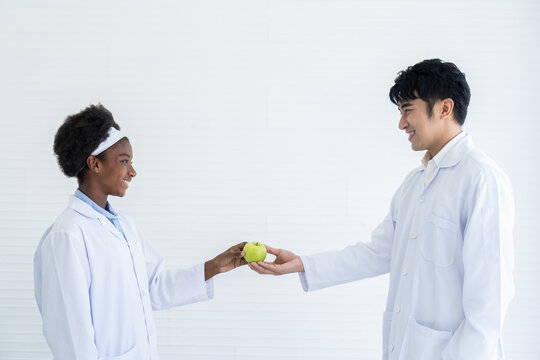 African American girl giving green apple to hand of Asian male science with white background. Happy diverse people giving green apple together - Powered by Adobe