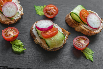 toasted baguette slices with cream cheese, cucumber, radish and tomato on a black slate board