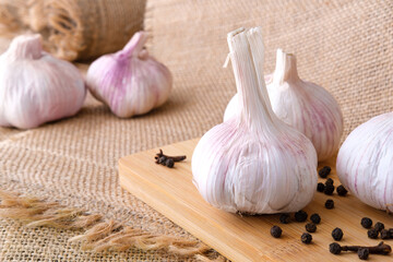 Garlic bulbs lie on a wooden cutting board. Black peppercorns and dried cloves are scattered all around. The table is covered with a rough burlap with fringe.