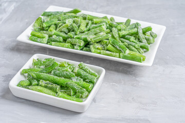 Selective focus of fresh frozen green beans vegetables covered with frost served in two rectangular plates preserving all vitamins and health benefits on gray concrete background. Horizontal image