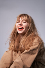 Attractive young fashion styled sensual woman laughing and posing on gray wall background dressed in hipster style beige jacket, with long hair and red lipstick