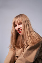 Sensual blonde woman laughing and posing on gray wall background dressed in hipster style beige jacket, with long hair and red lipstick. Fashion styled portrait