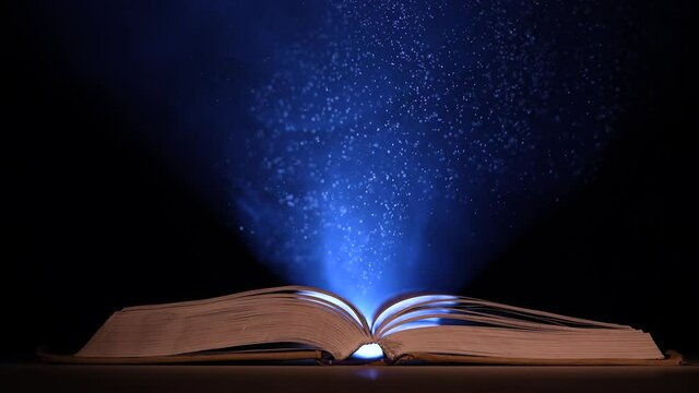In a dark, smoky studio, a thick, open book lies on a table against a black background. Particles of dust in the light of a searchlight fly up over an open book. Slow motion. Close up.