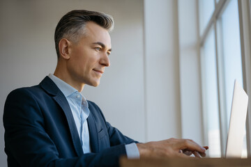 Confident mature businessman using laptop computer, typing, searching online, working in office  