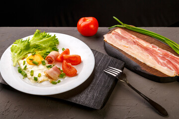 Scrambled eggs with bacon, garnished with tomato and green onion and on a gray concrete background on a wooden stand with a fork next to chopped bacon and tomato.
