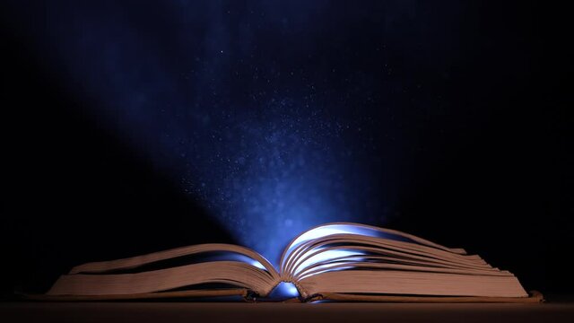 An open book falls on a table in a dark studio. A beam of light from a searchlight illuminates its pages and the dust particles that fly over it. Slow motion. Close up.