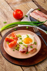 Scrambled eggs with bacon with tomato and green onion and on a wooden table in a plate with a fork next to chopped bacon and tomatoes.