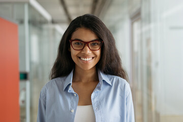 Young beautiful African American woman wearing stylish eyeglasses looking at camera, smiling. Portrait of confident manager standing in modern office 
