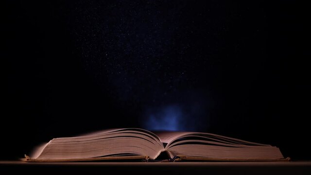 An open book falls on a table in a dark studio. A beam of light from a searchlight illuminates its pages and the dust particles that fly over it. Slow motion. Close up.