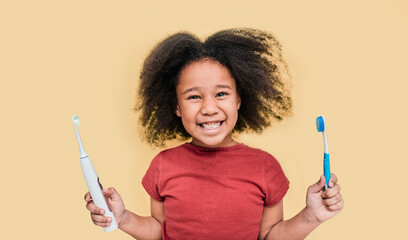 Smiling African American little girl holds manual and electric sonic toothbrushes. Child selects...